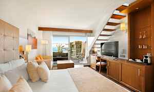 Et hotelværelse med trægulve, en hvid seng, en trappe og udsigt til en terrasse med hav i baggrunden. (AI-genereret tekst)