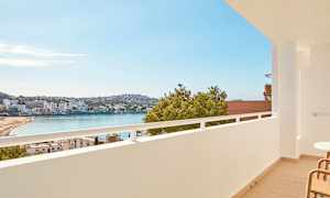 Udsigt fra en altan over en strand ved en bugt, med bygninger og grønt i baggrunden. (AI-genereret tekst)