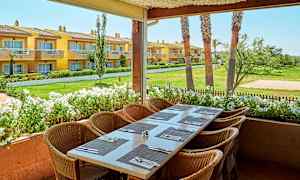 Billede af en udendørsterrasse med et dækket bord og stole, med udsigt over en have og gule bygninger i baggrunden. (AI-genereret tekst)