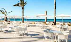 Udendørs siddeområde med hvide møbler og parasoller, samt palmer i baggrunden ved havet. (AI-genereret tekst)