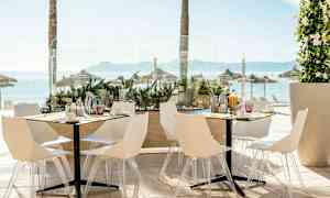 Billede af en udendørs restaurant med hvide stole og borde, med havudsigt og parasoller i baggrunden. (AI-genereret tekst)