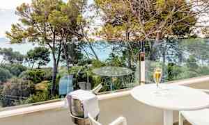 En balkon med udsigt over træer og havet. På balkonen står et hvidt bord med et glas champagne og en holder til en champagneflaske. (AI-genereret tekst)