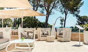 Siddeområde udendørs med hvide flettede møbler, beige puder og parasoller. Grønne træer og havet i baggrunden. (AI-genereret tekst)