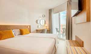 Et lyst hotelværelse med en stor seng, to gule puder, et skrivebordshjørne og en balkon med udsigt. (AI-genereret tekst)