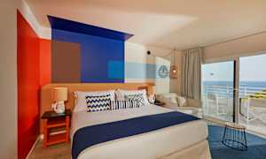 Et værelse med en dobbeltseng, dekorativt malede vægge, siddepladser og udsigt til havet gennem store vinduer. (AI-genereret tekst)