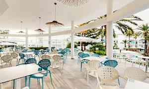 Udendørsterrasse med borde og stole, omgivet af palmer og hvide bygninger. (AI-genereret tekst)