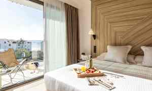 Et lyst hotelværelse med en redt seng med en morgenmadsbakke. Store glasdøre fører ud til en altan med udsigt over havet. (AI-genereret tekst)