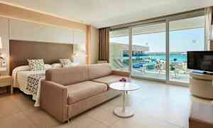 Hotelværelse med en seng, sofa, bord og udsigt til havet gennem glasdøre. (AI-genereret tekst)