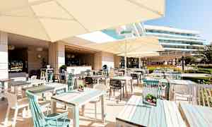 En udendørsrestaurant med borde og parasoller, omkringliggende bygninger ses i baggrunden. (AI-genereret tekst)