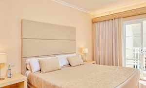 Værelse med en dobbeltseng, beige sengelinned og lampe på natbordet ved siden af vindue med gardiner. (AI-genereret tekst)