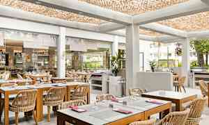 En lys restaurant med træborde og rattan-stole, placeret i en moderne indendørs atmosfære med et dekorativt loft og glasvægge, der åbner op mod udendørsområdet. (AI-genereret tekst)