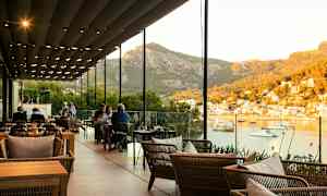 En restaurant med udendørs siddepladser under et overdække med udsigt over en havn og bjerge i baggrunden. (AI-genereret tekst)