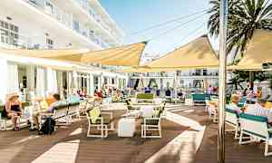 En udendørs terrasse ved en bygning med borde, stole og skygge-givende markiser. (AI-genereret tekst)