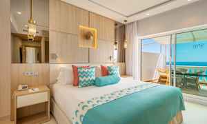 Hotelværelse med dobbeltseng, dekoreret i blåt og hvidt, med udsigt over havet fra en balkon. (AI-genereret tekst)