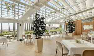 Restaurant med borde og stole under et glastag, omgivet af grønne planter og palmer. (AI-genereret tekst)