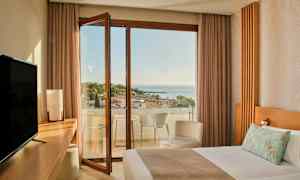 Et hotelværelse med en seng, skrivebord, TV og en åben altandør, der fører til en altan med udsigt over havet og træer. (AI-genereret tekst)