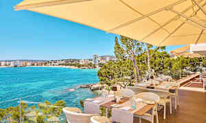 Udendørs servering med udsigt over en klarblå havbugt og bygninger i det fjerne. (AI-genereret tekst)