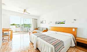 Et hotelværelse med lyst interiør, to enkeltsenge, en sofa, et bord og stole samt en balkon med udsigt. (AI-genereret tekst)