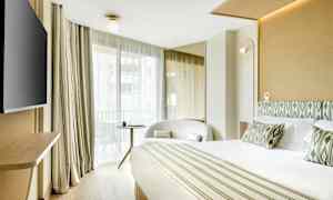 Et hotelværelse med en stor seng, lænestol, bord og et tv samt store vinduer med lyse gardiner. (AI-genereret tekst)