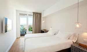 Et hotelværelse med to enkeltsenge, hvidt sengetøj, en væg-TV og udsigt over en by gennem glasdøre. (AI-genereret tekst)