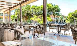 Udendørs siddeområde med borde og stole, omgivet af grønne træer og en bygning i baggrunden. (AI-genereret tekst)