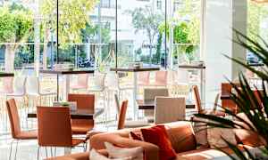 Et lyst og moderne siddeområde med borde og stole samt store vinduer, der giver udsigt til grønne områder udenfor. (AI-genereret tekst)