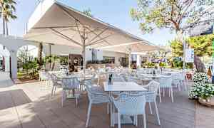 En udendørsrestaurant med hvide borde og stole samt store parasoller. (AI-genereret tekst)