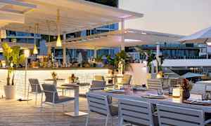 En udendørsrestaurant med hvide møbler og grønne planter, beliggende nær en bygning med glasvinduer. (AI-genereret tekst)