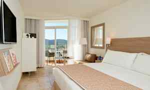 Et hotelværelse med en stor seng, siddeområde og adgang til en balkon med udsigt over træer og landskab. (AI-genereret tekst)