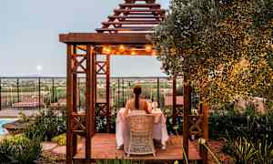 En person sidder ved et bord under en træpergola dekoreret med lys, omgivet af beplantning med udsigt over landskabet. (AI-genereret tekst)