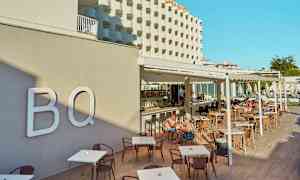 Hotellets facade og udendørs serveringsområde med borde og stole, mennesker der sidder og spiser. (AI-genereret tekst)