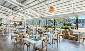 Udendørs servering ved havet med borde og stole samt udsigt til både i havnen. (AI-genereret tekst)