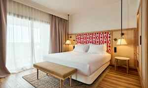 Et hotelværelse med en dobbeltseng, lyse gardiner og en bænk foran sengen. (AI-genereret tekst)