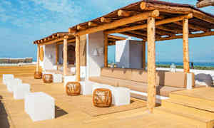 Terrasse med solbeskyttelse af træ, siddeområder, hvide kubeformede borde og udsigt over havet. (AI-genereret tekst)