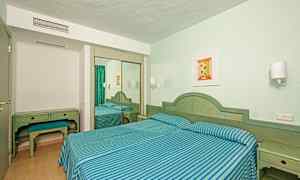 Et hotelværelse med to enkeltsenge dækket med blå- og grønstribede sengesæt, skrivebord med skammel og et spejlklædeskab. (AI-genereret tekst)