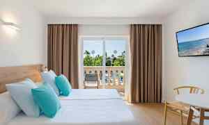 Et hotelværelse med to enkeltsenge med lyseblå og turkise puder, en balkon med udsigt over haven, og et TV på væggen. (AI-genereret tekst)