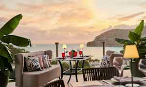 Udendørs servering med borde og sofaer, omgivet af grøn beplantning, med udsigt over havet under en solnedgang. (AI-genereret tekst)