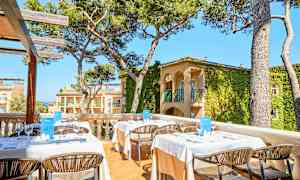 Billede af en udendørs terrasse med opdækkede borde og stole, omgivet af træer og bygninger med plantebeklædte facader. (AI-genereret tekst)