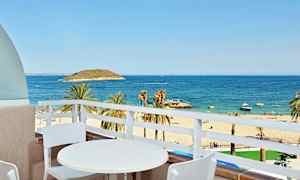 Udsigt fra en balkon med et hvidt bord og to stole mod en sandstrand, palmer og et blåt hav. (AI-genereret tekst)