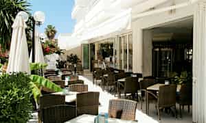 Udendørsservering med mørke rottingstole og hvide borde, omgivet af grønne planter. (AI-genereret tekst)