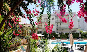 En bygning med flere altaner bag hængende lyserøde blomster, en swimmingpool og liggestole. (AI-genereret tekst)