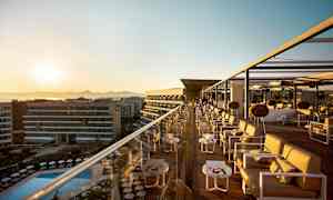 En tagterrassebar med siddemøbler og borde ved solnedgang, med udsigt over en pool og bygninger. (AI-genereret tekst)