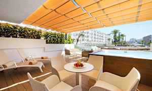 En udendørs terrasse med liggestole, borde og stole. Der er en lille pool og udsigt til hotelbygninger. (AI-genereret tekst)