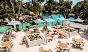 En terrasse med borde, stole og parasoller nær en turkis vandstrand og grønne områder. (AI-genereret tekst)