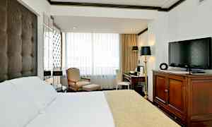 Et hotelværelse med en seng med hvidt og beige sengetæppe, en lænestol og skammel ved vinduet, et skrivebord med stol og et TV placeret på en trækommode. (AI-genereret tekst)