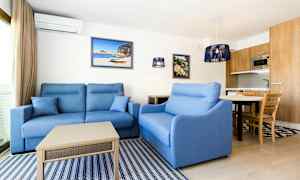 En stue med blå sofaer, et sofabord, væghængte billeder og et køkken med træelementer i baggrunden. (AI-genereret tekst)