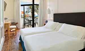 Et lyst hotelværelse med to enkeltsenge, et skrivebord og en balkon med udsigt over havet og palmer. (AI-genereret tekst)