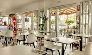 En restaurant med hvide stole og borde, åben ud mod en terrasse og omkringliggende grønne områder. (AI-genereret tekst)