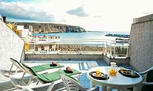 Terrasse med to solvogne og et bord med appelsinjuice og bagværk, havudsigt i baggrunden. (AI-genereret tekst)