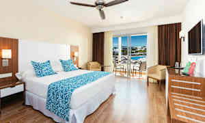 Et hotelværelse med en stor seng, blå dekorative puder og tæppe samt et bord med stole ved en glasdør, der fører til en balkon. (AI-genereret tekst)